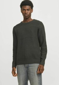 Maglione a maglia testurizzato verde scuro con scollo rotondo e polsini a coste, abbinato a jeans azzurri chiari. Vestibilità classica, design casual.