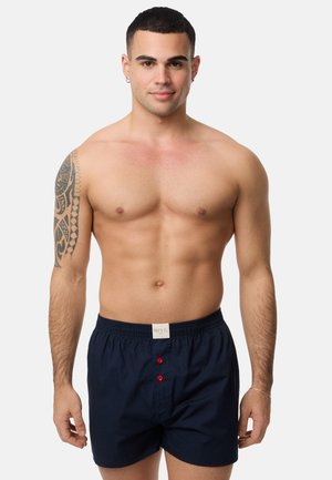 Boxers de algodón azul marino con una cinturilla elástica, con botones rojos en la parte delantera y una etiqueta en la cintura. Sin patrones ni diseños adicionales.