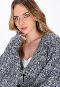 Cardigan grigio a maglia con vestibilità ampia, dotato di un davanti annodato e di un motivo testurizzato. Materiale morbido e peloso con sfumature di blu.