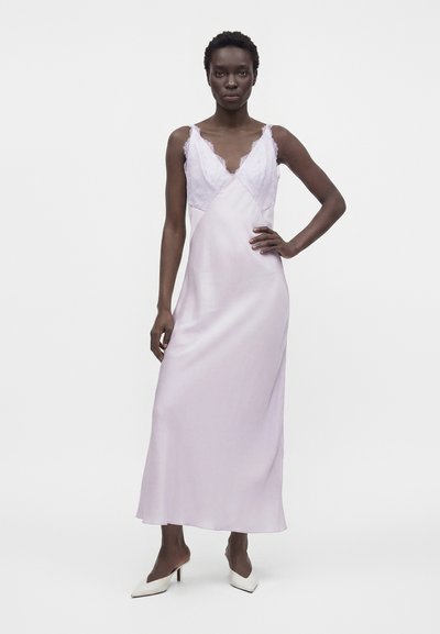 Femme portant une robe nuisette en satin lavande clair avec des détails en dentelle, debout avec une main sur la hanche et des escarpins blancs à bout pointu sur fond uni.