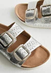 Spännbandssandaler med metallic silverfinish, perforerat mönster på remmarna, bruna mockainlägg och strukturerade gummisulor.