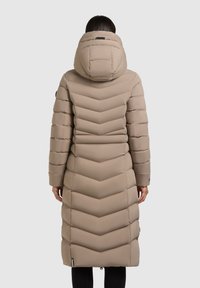 Beige hosszú puffermellény chevron steppeléssel, állítható kapucnival és diszkrét logóval. Jellemzői az elasztikus mandzsetta és az oldalsó cipzáros zsebek.