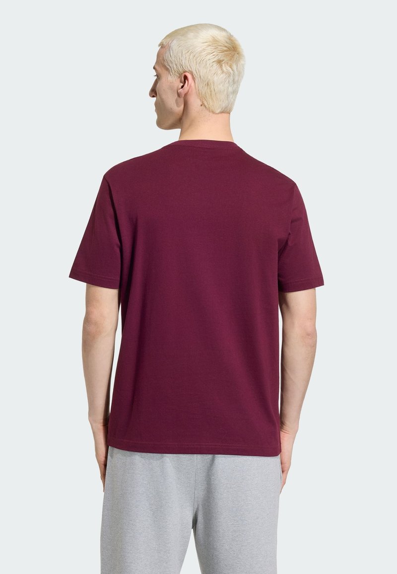 Jeune homme aux cheveux blonds courts portant un t-shirt bordeaux uni à manches courtes et un pantalon gris clair, vu de dos.