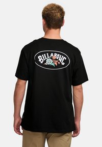 Melna kokvilnas T-krekls ar lielu, daudzkrāsainu ziedu logo un "BILLABONG" tekstu uz muguras. Īsas piedurknes un brīvi pieguļoša silueta.