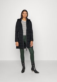Doudoune noire avec capuche, texture matelassée, fermeture éclair à l'avant, associée à un haut rayé et un pantalon vert, complétée par des bottes à talons noires.
