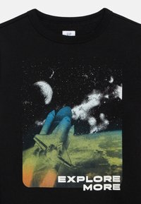 Διαστημικό λεωφορείο που εκτοξεύεται πάνω από τη Γη με αστέρια, το φεγγάρι και τις λέξεις «ΑΝΑΚΑΛΥΨΕ ΠΕΡΙΣΣΟΤΕΡΑ» σε μαύρο μπλουζάκι.