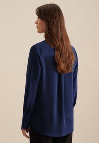 Blusa a maniche lunghe blu navy con texture liscia, polsini con bottoni e orlo curvo. Presenta un colletto classico e un rinforzo sulle spalle.