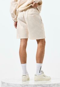 Beige Cargo-Shorts aus Baumwolle mit lockerer Passform, ausgestattet mit Seiten- und Gesäßtaschen, kombiniert mit weißen Socken und cremefarbenen Reebok-Schuhen.
