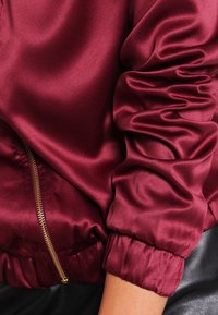 Burgundy satin bomber jacket with a gold zipper. Features gathered sleeves and elastic cuffs for a fitted look. Smooth texture.