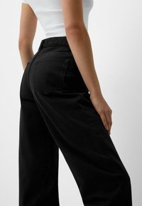 Jeans anchos negros de tiro alto, con dos bolsillos traseros y una textura suave y mate. Combinándolos con una blusa blanca ajustada.