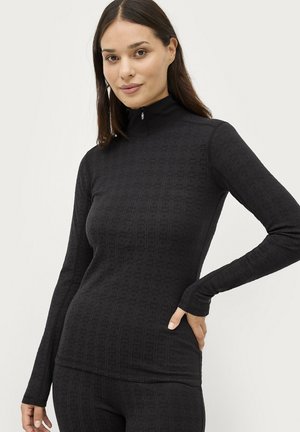 Kvinde iført sort mønstret langærmet top med høj krave og delvis lynlås, poserer med den ene hånd på hoften foran en ensfarvet baggrund.