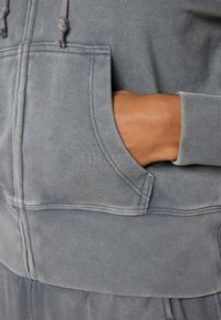 Hoodie gris avec une poche avant, des poignets côtelés et des cordons de serrage. Tissu doux et texturé avec des détails de couture visibles.