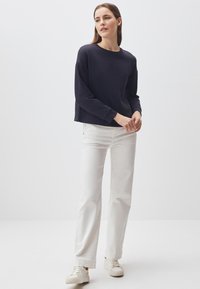 Felpa scura a maniche lunghe, con scollo rotondo, abbinata a jeans bianchi a gamba dritta e sneakers bianche su uno sfondo neutro.