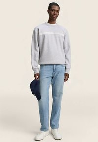 Grijze sweatshirt met een witte horizontale streep, lichtblauwe spijkerbroek, witte sneakers en een marineblauwe baseballpet in de linkerhand.