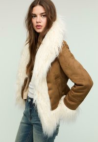 Brun faux suede jakke med hvid faux pels krave, med en tætsiddende design og sidelommer, båret over blå jeans.