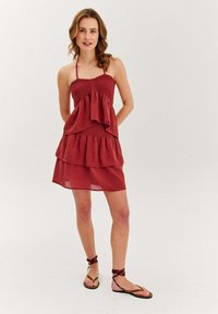 Žena v bezrukávových červených šatách s volánmi, skombinovaných s hnedými sandálmi s viazaním okolo členkov, stojí na neutrálnom pozadí.