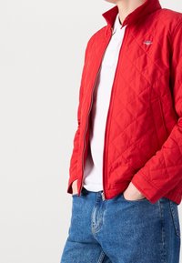 Giacca rossa trapuntata con colletto alto, chiusura con zip e tasche laterali, indossata sopra una camicia bianca e jeans in denim blu.