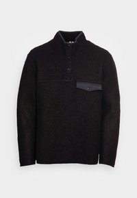 Mustapullovari, joka on valmistettu teksturoidusta kankaasta, korkealla kauluksella, kolmella mustalla painikkeella ja etupuolen rintataskulla, jossa on läppä ja painikekuin.