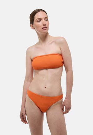 Femme à la peau claire et aux cheveux bruns portant un haut de bikini bandeau orange texturé et un bas de bikini assorti, debout devant un fond uni.