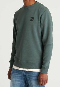 Groene sweatshirt met textuur, ribstof, ronde hals en een klein zwart logo op de borst. Gecombineerd met lichtblauwe jeans.