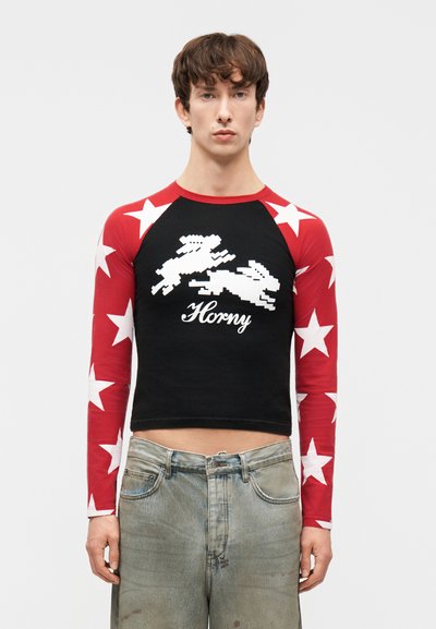 Mladý muž nosiaci čierny crop top s dlhými rukávmi s červeným vzorom hviezd, pixelovanými zajacmi a nápisom „Horny“ na hrudi.