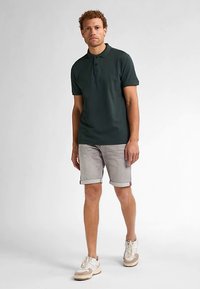 Tummanvihreä lyhythihainen poolopaita, yhdistetty vaaleanharmaisiin farkkushortseihin ja valkoisiin tennareihin, joissa on beiget yksityiskohdat, seisoo neutraalilla taustalla.