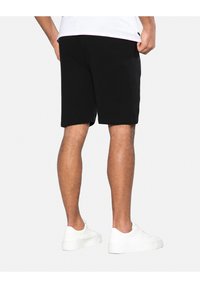 Shorts en coton noir avec une taille élastique, arrivant au milieu de la cuisse, associés à des baskets blanches. Texture lisse, design simple, sans motifs visibles.