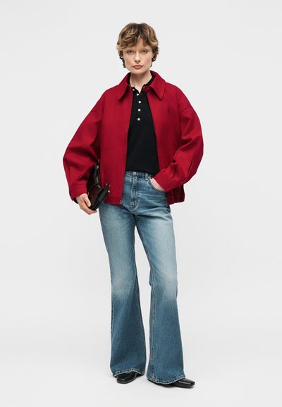 Γυναίκα που φορά ένα κόκκινο μπουφάν με φερμουάρ, μαύρο πόλο πουκάμισο, φαρδύ μπλε τζιν, μαύρα παπούτσια, κρατώντας μια μαύρη τσάντα χειρός, στέκεται μπροστά σε απλό φόντο.