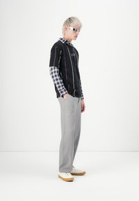 Joven con cabello rubio platino, vistiendo una camiseta negra de rayas en capas sobre una camisa de cuadros azul, pantalones grises, zapatillas blancas y gafas de sol blancas.