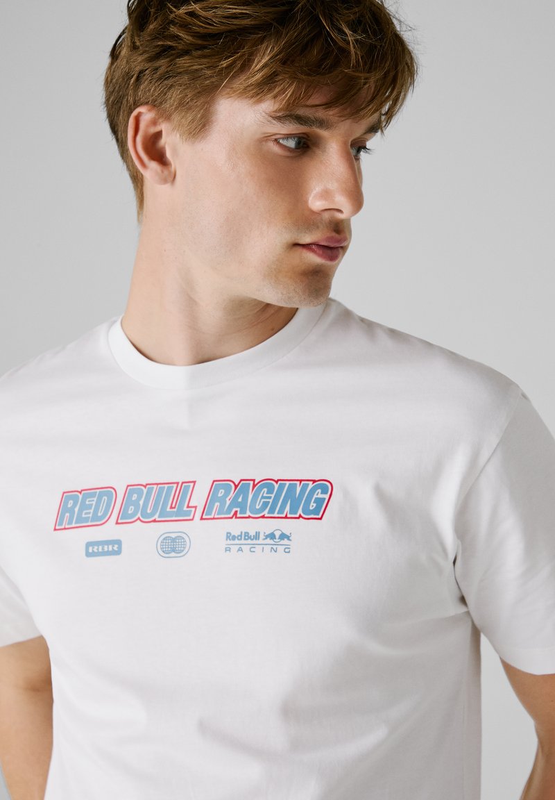 Biała koszulka z bawełny, z centralnym nadrukiem w niebieskim i czerwonym kolorze, z napisem "RED BULL RACING" oraz logo. Design z okrągłym dekoltem.