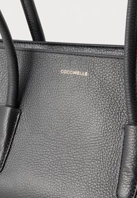 Borsa in pelle nera con texture, dotata di due manici e del marchio COCCINELLE in oro sulla pannello frontale.