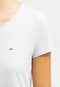 Bílá krátkosrstá tričko s kulatým výstřihem a malým vyšitým logem v modré, červené a bílé barvě na levé části hrudi.