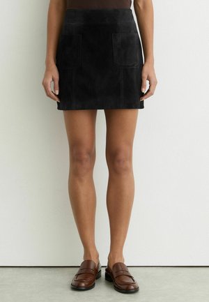 Žena v čiernej semišovej mini sukni s prednými nášivkovými vreckami, hnedých mokasínach a tmavohnedom topu stojaca pri bielej stene.