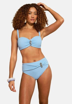 Frau mit lockigem Haar, die einen hellblauen, strukturierten Bikini mit Seepferdchen-Broschen trägt, posiert mit einer Hand am Kopf und Armbändern am Handgelenk.