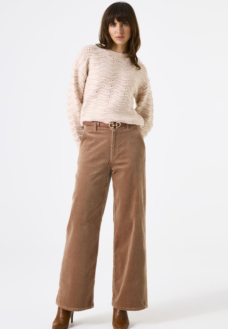 Beige gebreide trui met golvenpatroon, gecombineerd met een bruine corduroy met brede pijpen en een bruine riem met een ronde gesp. Bruine haklaarzen.
