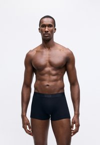 Fit man standing against plain background wearing black Hugo boxer briefs, facing forward with a neutral expression.