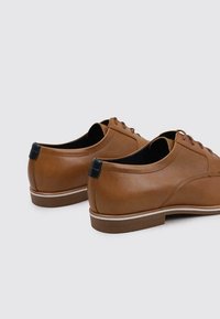 Chaussures en cuir marron avec une texture lisse, bout rond et un capuchon de talon foncé contrastant. Elles présentent un design à lacets et une semelle blanche subtile.