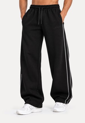 Modelo con pantalones jogger negros de pernera ancha, cintura con cordón y ribete blanco en los laterales, combinados con zapatillas blancas.