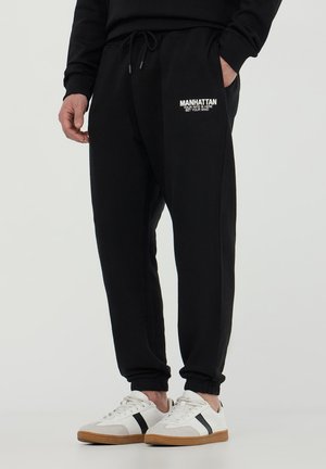 Pantaloni jogger neri con polsini elasticizzati, dotati di vita con cordino, tasche laterali e testo bianco "MANHATTAN" sulla gamba sinistra.