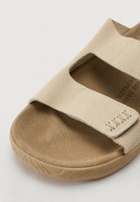 Beige mockasinsandal med två breda remmar, rund tå, texturerad fotbädd och gummisulor med ett subtilt mönster.