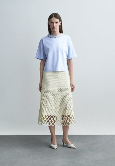 Γυναίκα με ανοιχτό μπλε τοπ κοντών μανικιών με διακοσμημένο ντεκολτέ, μπεζ μίντι φούστα με σχέδιο πλέγματος και λευκά μυτερά τακούνια.