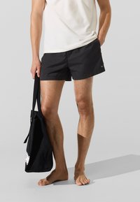 Čierne plavkové šortky z ľahkého materiálu s plochým pásom a jemným logom značky. Držané vedľa čiernej tašky na rameno.