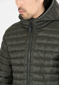 Olivgrön puffjacka med hög krage, horisontellt quiltad textur, hel dragkedja fram och justerbar huva. Lättviktsmaterial.