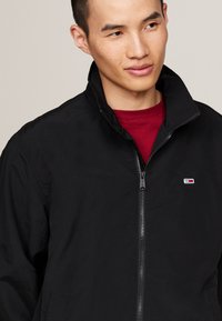 Zwarte ritsjas van lichtgewicht stof; heeft een staande kraag en een klein merklogo op de linkerkant van de borst. Draag het over een rood shirt.