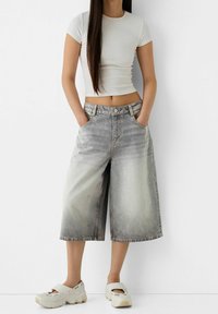 Shorts en denim gris clair coupés, au design délavé, avec des poches latérales et une fermeture à bouton. Associés à un haut ajusté blanc et des chaussures beiges.