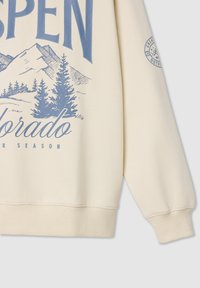 Beżowy sweatshirt z napisem "ASPEN" w niebieskim kolorze, grafiką gór, zielonymi drzewami iglastymi oraz prążkowanymi mankietami i dołem.