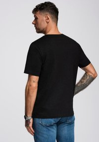 Černé tričko s krátkým rukávem vyrobené z bavlny, s kulatým výstřihem a hladkou strukturou, nošené s modrými džíny. Viditelné tetování na paži.