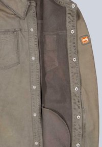 Olivgrüne Jacke mit Druckknöpfen, zwei Fronttaschen und einem strukturierten Innenfutter. Sie hat Kontrastnähte und ein kleines Logotag.