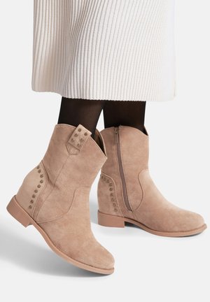 Botines beige de ante con cremallera lateral y detalles de tachuelas, combinados con medias negras y una falda blanca de canalé.