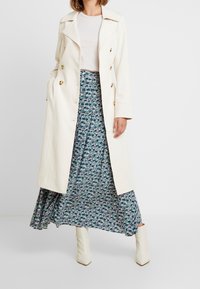 Langer cremefarbener Mantel mit großen goldenen Knöpfen, kombiniert mit einem blumengemusterten blauen Rock. Die Stiefel sind weiß mit einem quadratischen Zehendesign.